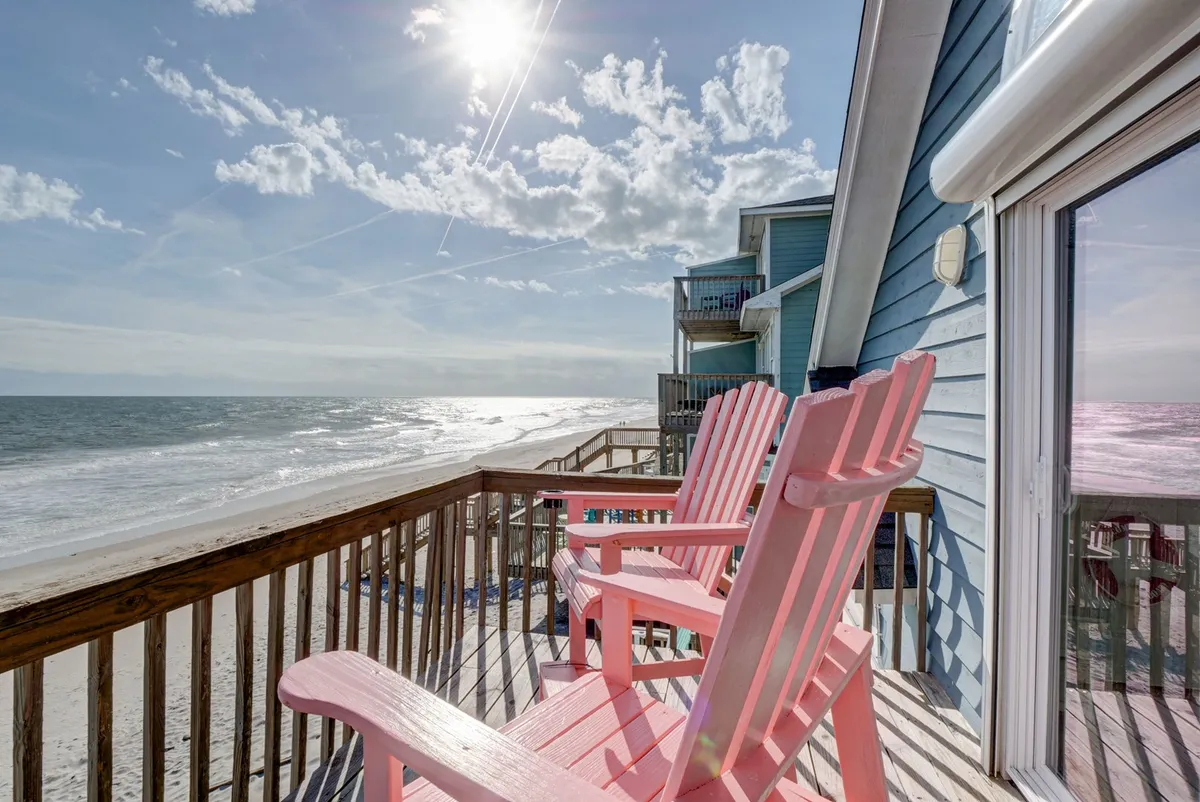 The image showcases a pair of vibrant pink Adirondack chairs facing the sparkling ocean, bathed in warm sunlight. The tranquil setting is perfect for soaking in sunrise views and enjoying the soothing sounds of the waves, with the expansive shoreline stretching into the distance.