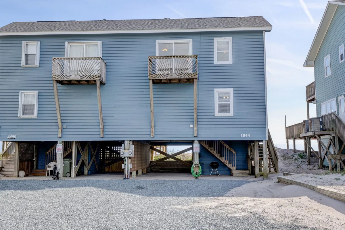 This exterior shot captures the vacation home with its two spacious balconies, blue siding, and an inviting underneath space that hints at direct beach access. The scene suggests areas perfect for gathering, playing games, or simply relaxing outside after a day of exploration.