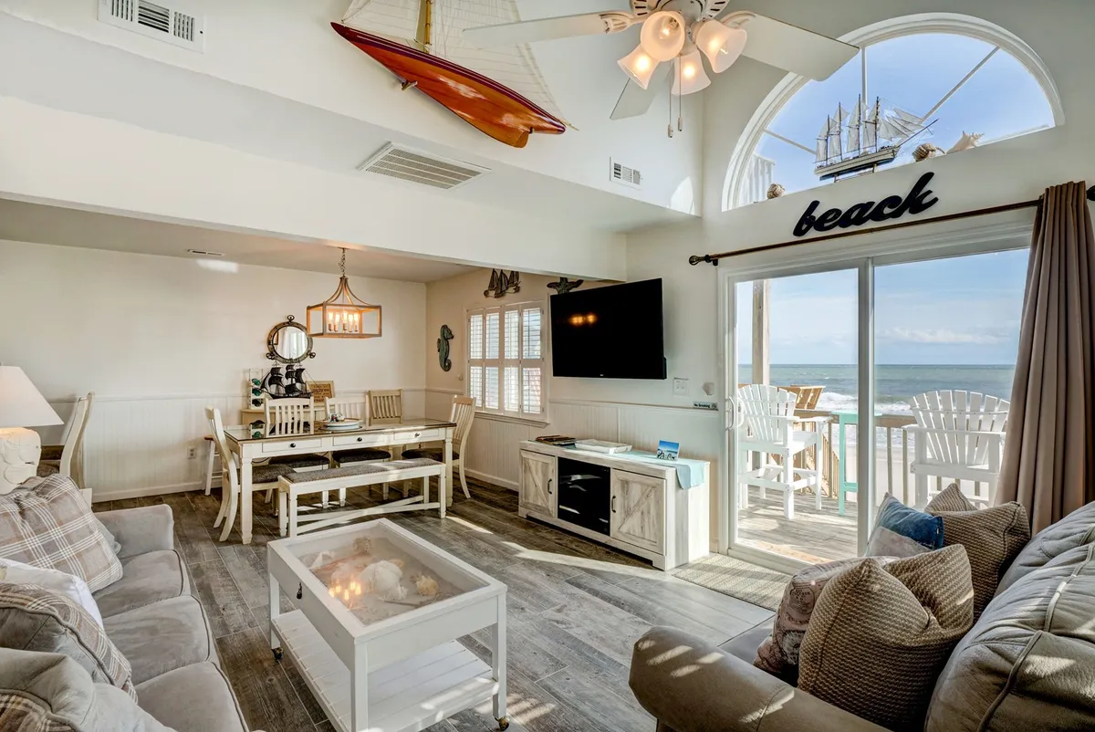 A bright, open living and dining area featuring a comfortable sofa, a sizable dining table, a large TV, and expansive windows offering sweeping ocean views. Decorative elements like a model sailboat add a coastal feel, and the room seamlessly connects relaxation, entertainment, and communal meals.