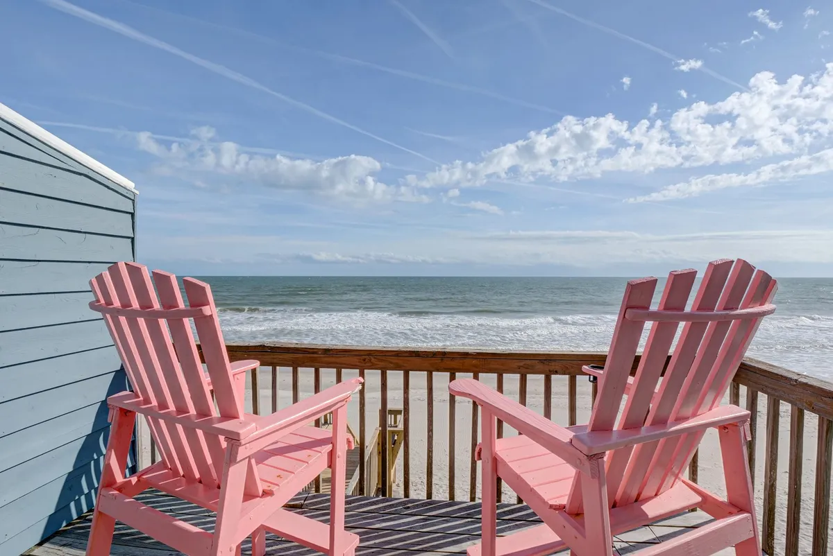 A set of pink adirondack chairs is arranged on a deck with unobstructed views of the ocean and blue sky. The scene radiates calm and comfort, providing a serene spot for morning coffee or sunset watching after playtime on the beach. The openness suggests a space where everyone—pets included—can unwind.