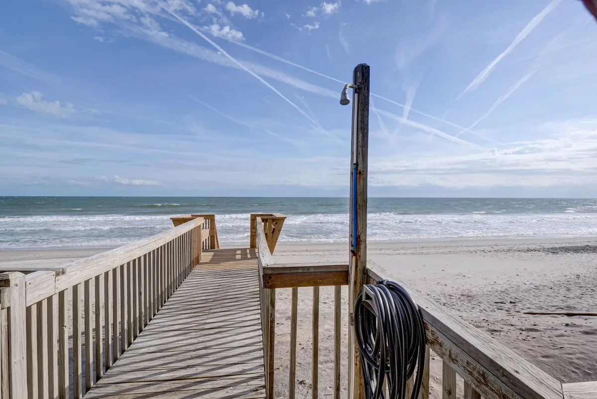 A wooden walkway extends toward the sparkling ocean, with a convenient outdoor shower post at the start of the sandy beach area. The scene is bright and inviting, emphasizing the easy transition between property and coastline.