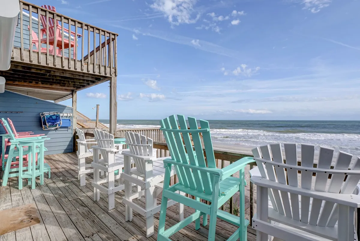 This exterior image showcases a set of colorful Adirondack chairs lined up on a private deck, with a sweeping ocean view in the background. The scene is inviting and tranquil, with the deck directly overlooking the sandy beach, emphasizing the seamless connection between the home and the sea.