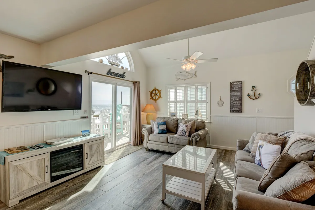 A cozy living room set-up with plush sofas facing a large flat-screen TV, a light coffee table, and a window framing ocean views. The space invites family movie nights, relaxed conversations, and provides a glimpse of the outdoors, reinforcing the indoor-outdoor lifestyle.