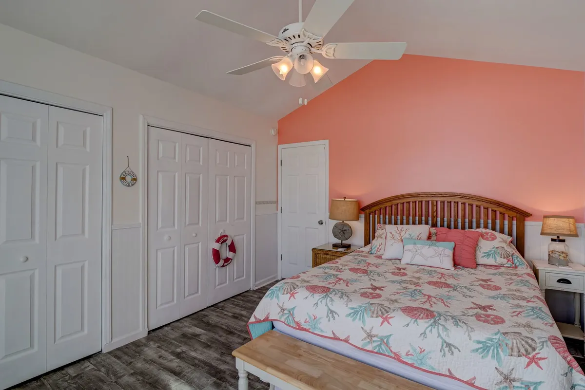 This bedroom features colorful bedding, a vibrant accent wall, nightstands with lamps, a ceiling fan, and a wooden bench at the foot of the bed. The room is bathed in warm sunlight, creating a cozy and cheerful ambiance.