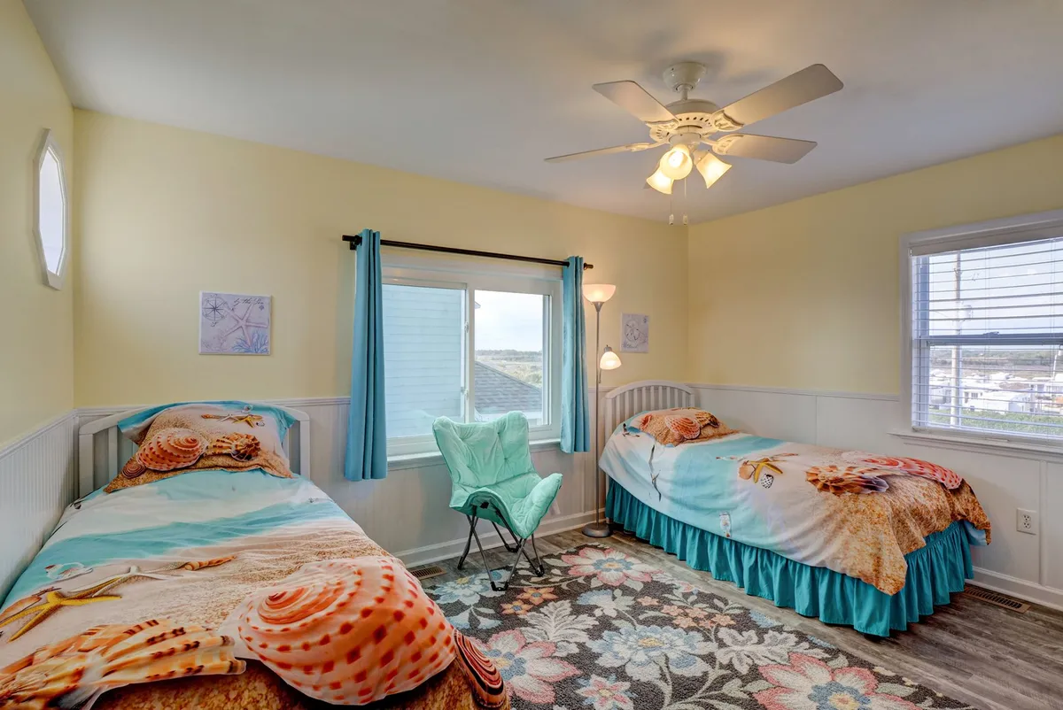 A playful and inviting twin bedroom with two beds featuring beach-themed bedding, a large window that lets in ample natural light, and a colorful area rug. The room feels vibrant and perfect for children or friends.