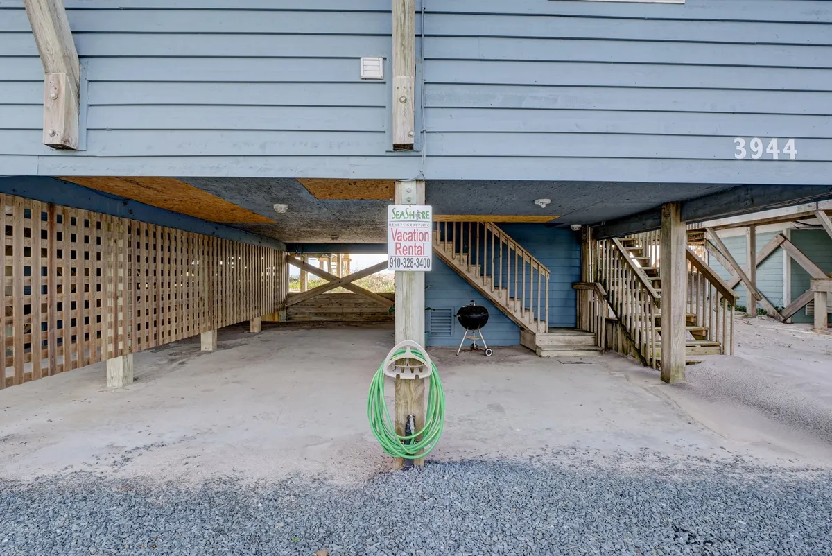 The image showcases the exterior of the beachfront duplex, highlighting sturdy wooden pillars, a vacation rental sign, a green garden hose, and a grill. The outdoor area invites guests to enjoy barbecuing, relaxing in the shade, and dining outdoors, with a relaxed and casual mood that suits year-round enjoyment.