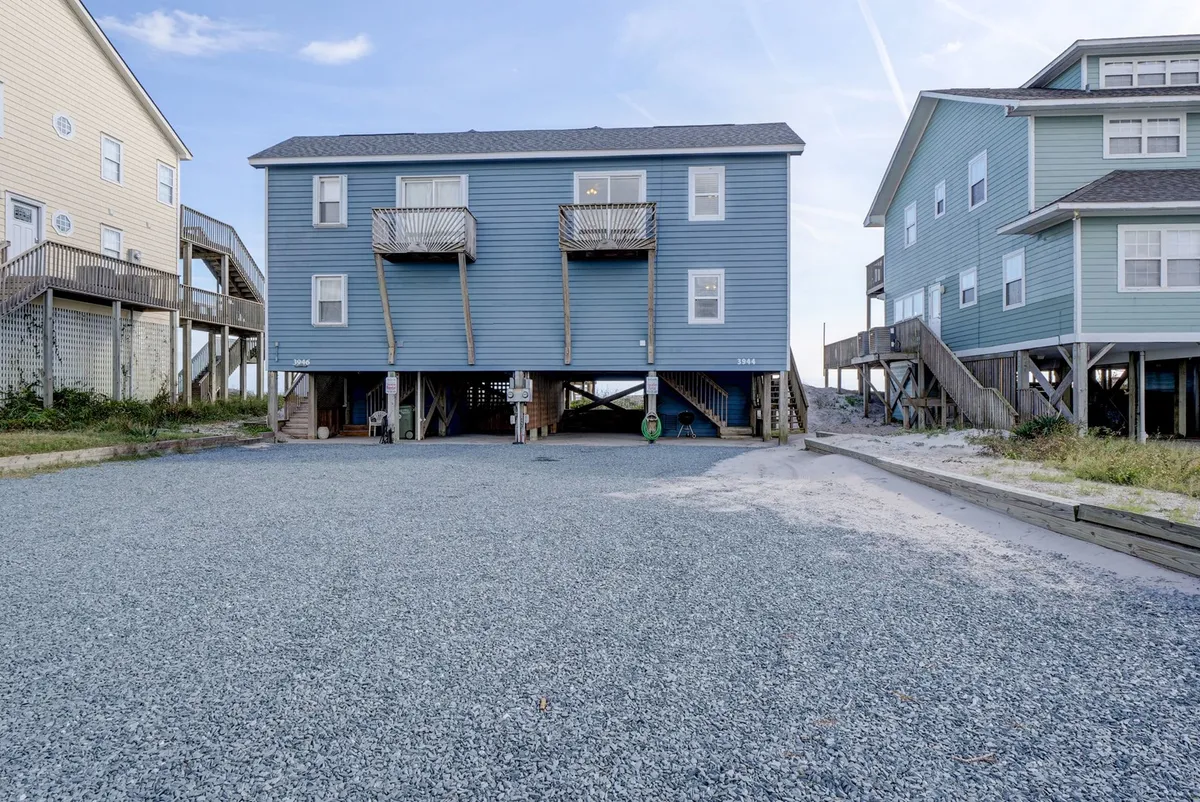 A wide shot of the blue vacation home exterior, featuring a gravel parking area and lush surrounding vegetation. The home feels inviting and close to nature, suggesting ample space and easy access for guests with pets.
