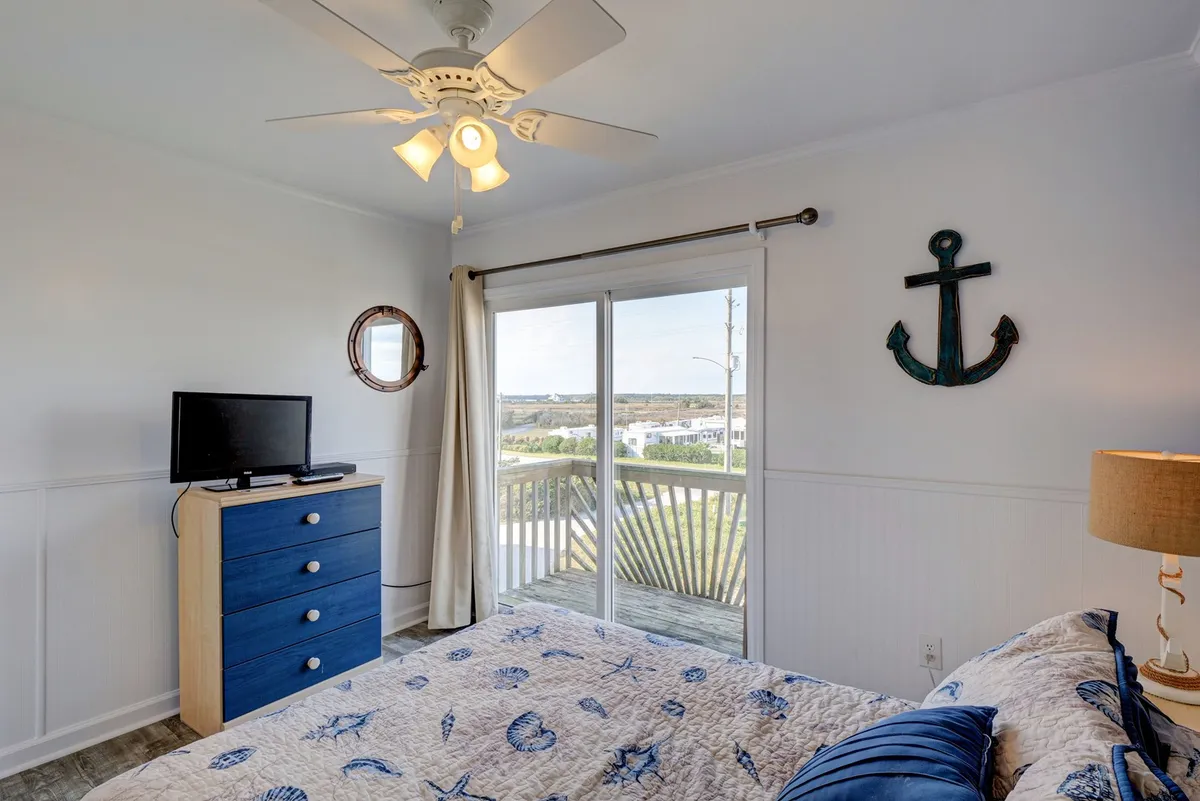 This image showcases a bedroom with nautical-themed bedding, a blue dresser, a television, anchor wall decor, and sliding glass doors that open to a private balcony. The balcony offers ocean views, and the space is filled with natural light, creating a tranquil and refreshing atmosphere.