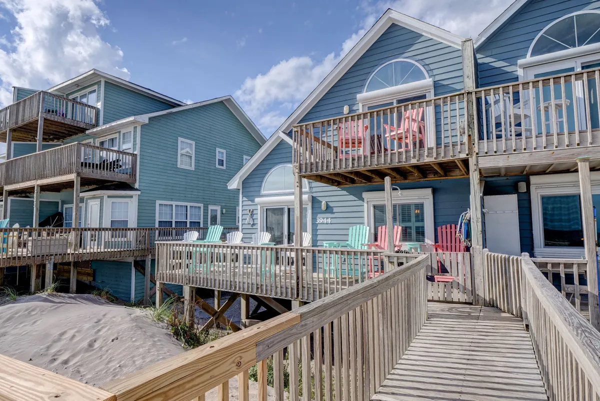 The image shows a breezy deck area with colorful adirondack chairs, wooden walkways, and direct beachfront access. The deck overlooks the sand and sea, inviting guests to relax, socialize, and soak up the coastal atmosphere. The setup suggests plenty of space for both children and pets to play while adults unwind nearby.